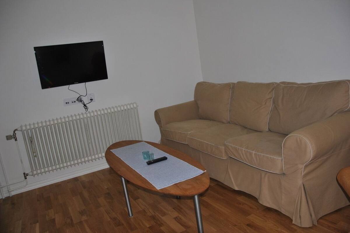 A living room with a beige sofa, a wooden coffee table with a remote control, a flat-screen TV mounted on the wall, and a white radiator.