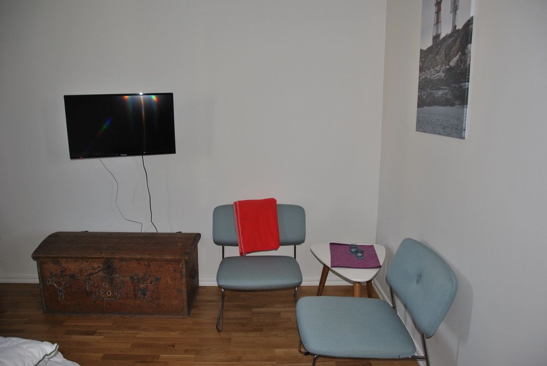A room with a television mounted on the wall, a wooden chest, two chairs, a small table with a purple cloth, and a framed picture on the wall.