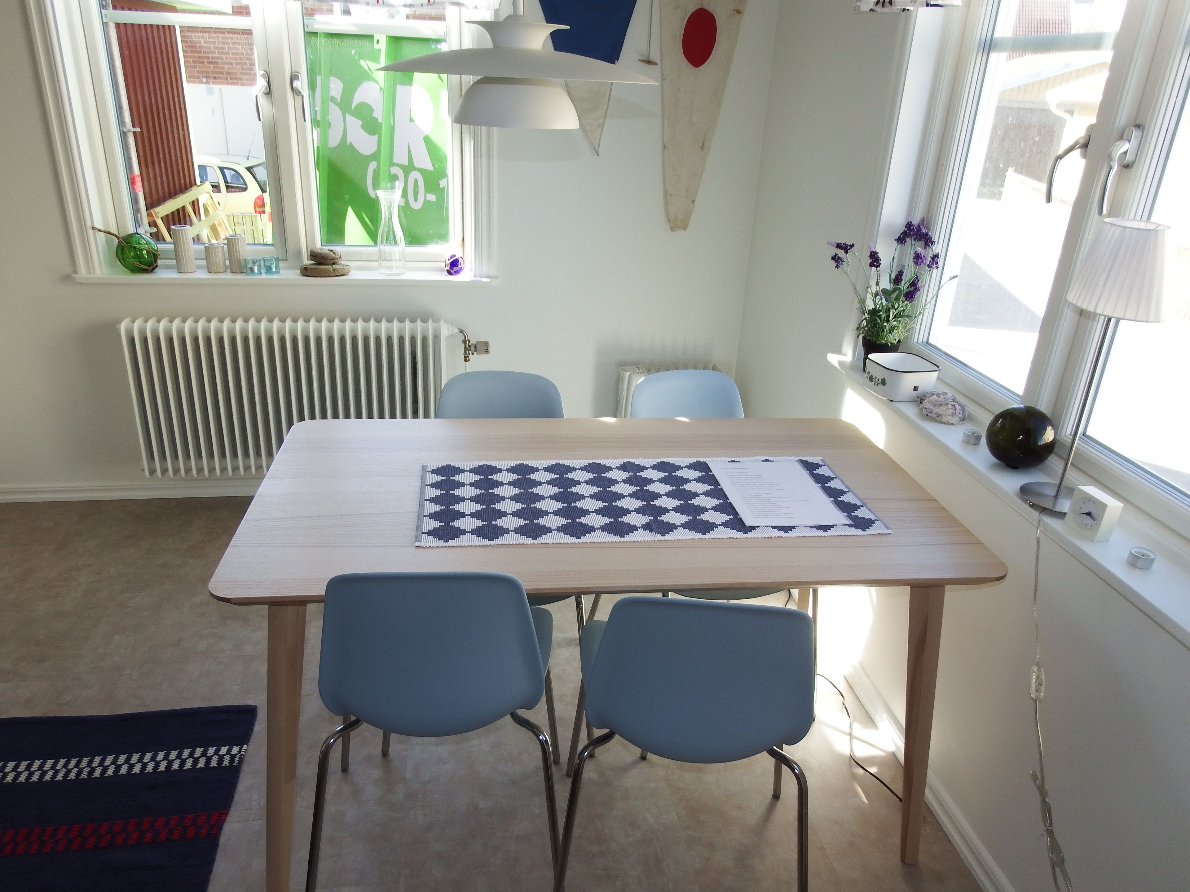 Una habitación con una mesa de madera, dos sillas azules, un radiador, un ventanal con vistas al exterior y un tablero de ajedrez sobre la mesa.