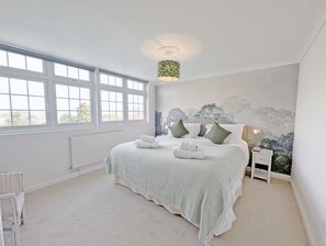 Une chambre avec une grande fenêtre, un lit recouvert d’une couvre-lit à motifs, une table de chevet avec une lampe et une fresque murale.