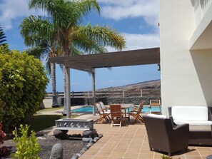 Relax in a serene patio setting with ocean and mountain views, surrounded by a pergola and lush greenery.