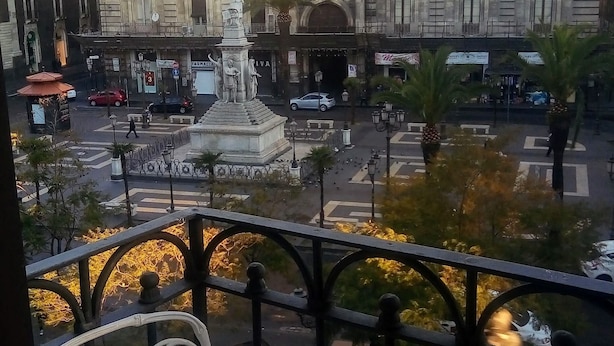 Un balcón con barandilla de metal blanco ornamentada y una silla blanca. Al fondo, un edificio grande con arquitectura clásica, una estatua y una calle concurrida con coches y peatones.