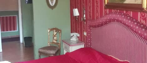 A room with a large bed, a chair, a bedside table with a lamp, a framed picture on the wall, and a door leading to another room.