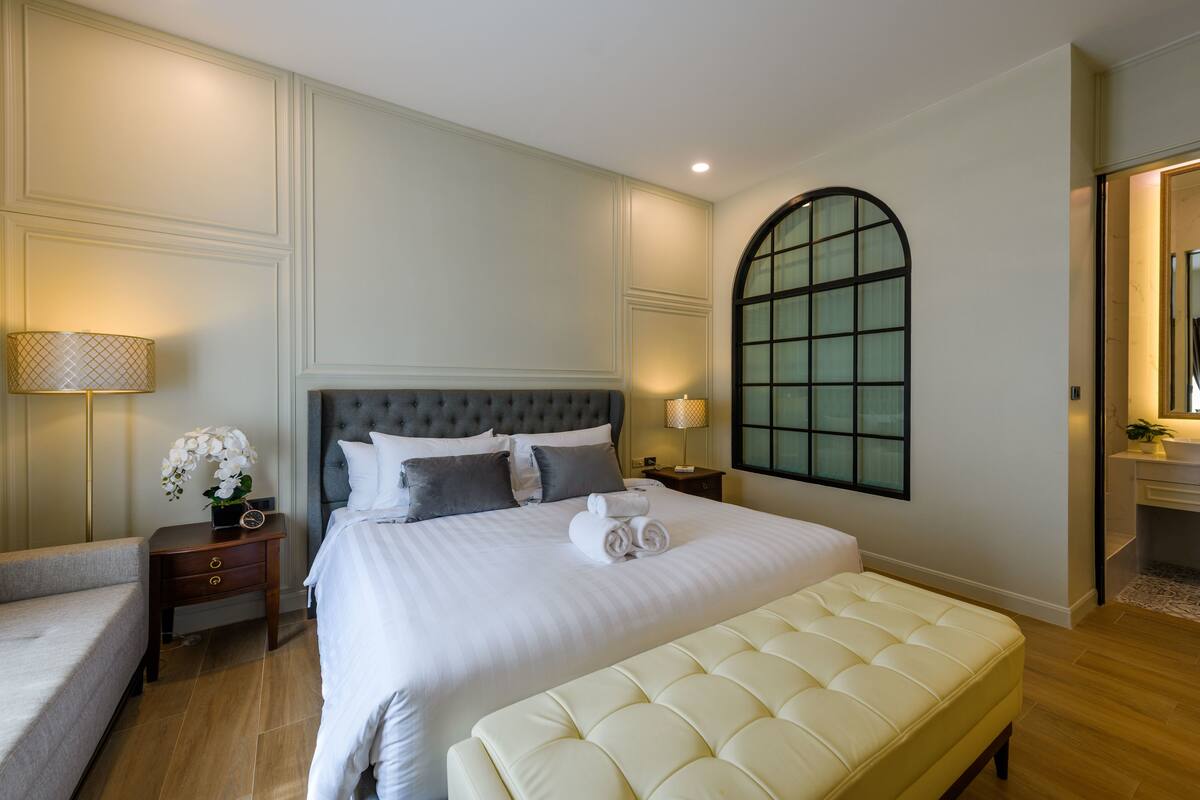 Un dormitorio ordenado con una cama grande, una mesita de noche con lámpara, un banco y un amplio ventanal de arco.