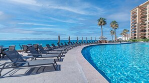 A resort pool with lounge chairs, palm trees, and a view of the ocean.