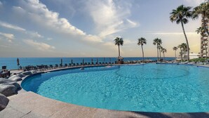 Una piscina curvada con agua azul cristalina, rodeada de sillones de descanso y palmeras, con vistas al mar tranquilo.