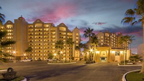 Un hotel de varios pisos con un patio central, palmeras y un cielo al atardecer.