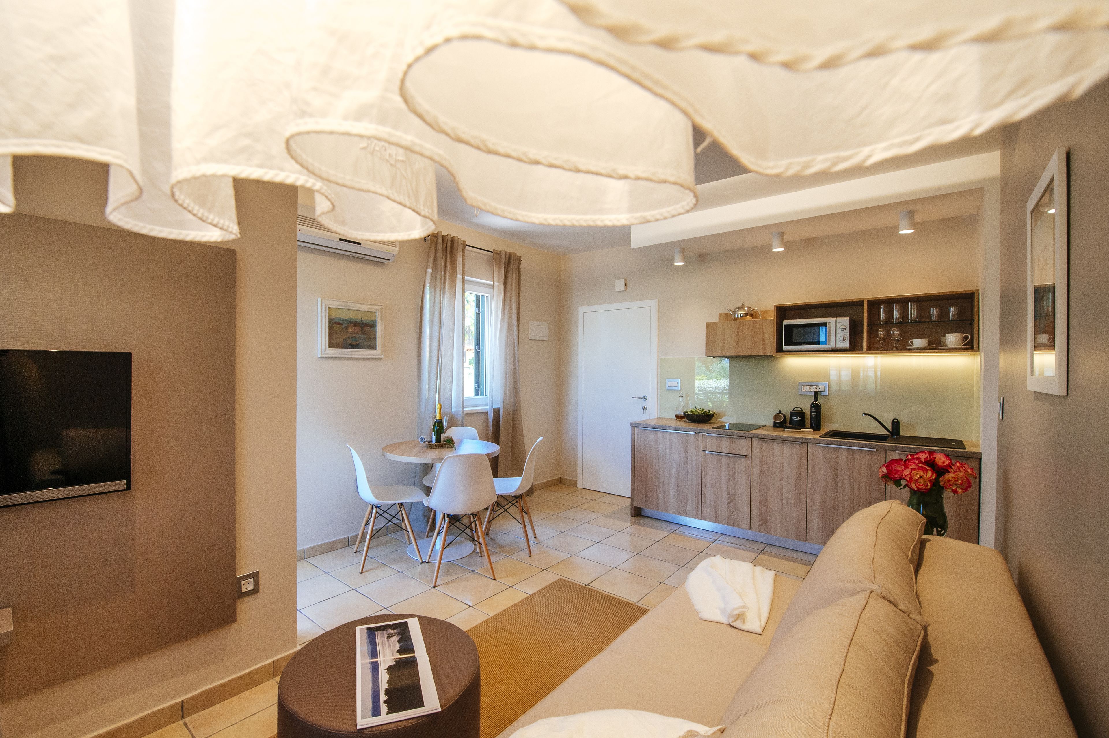 A modern living room with a sofa, a dining table with chairs, a kitchen area with a microwave, and a TV mounted on the wall.