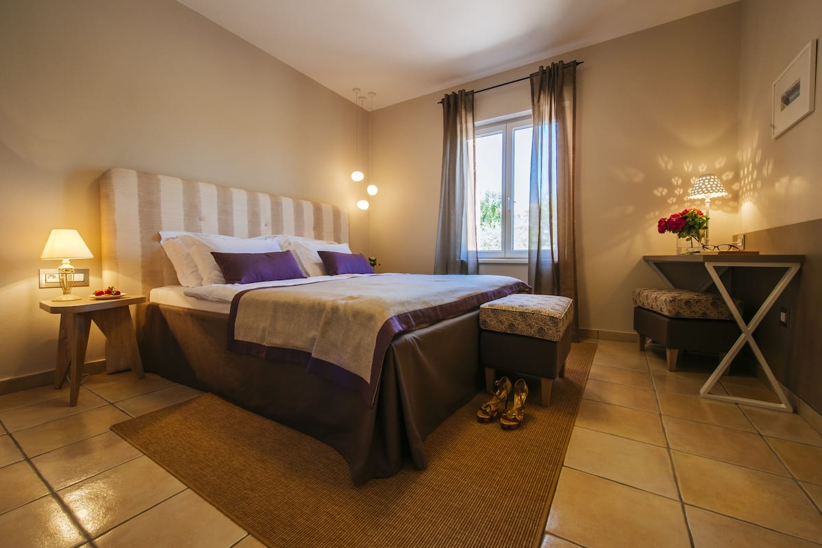 A neatly arranged bedroom with a bed, bedside tables, a window with curtains, and a small seating area.