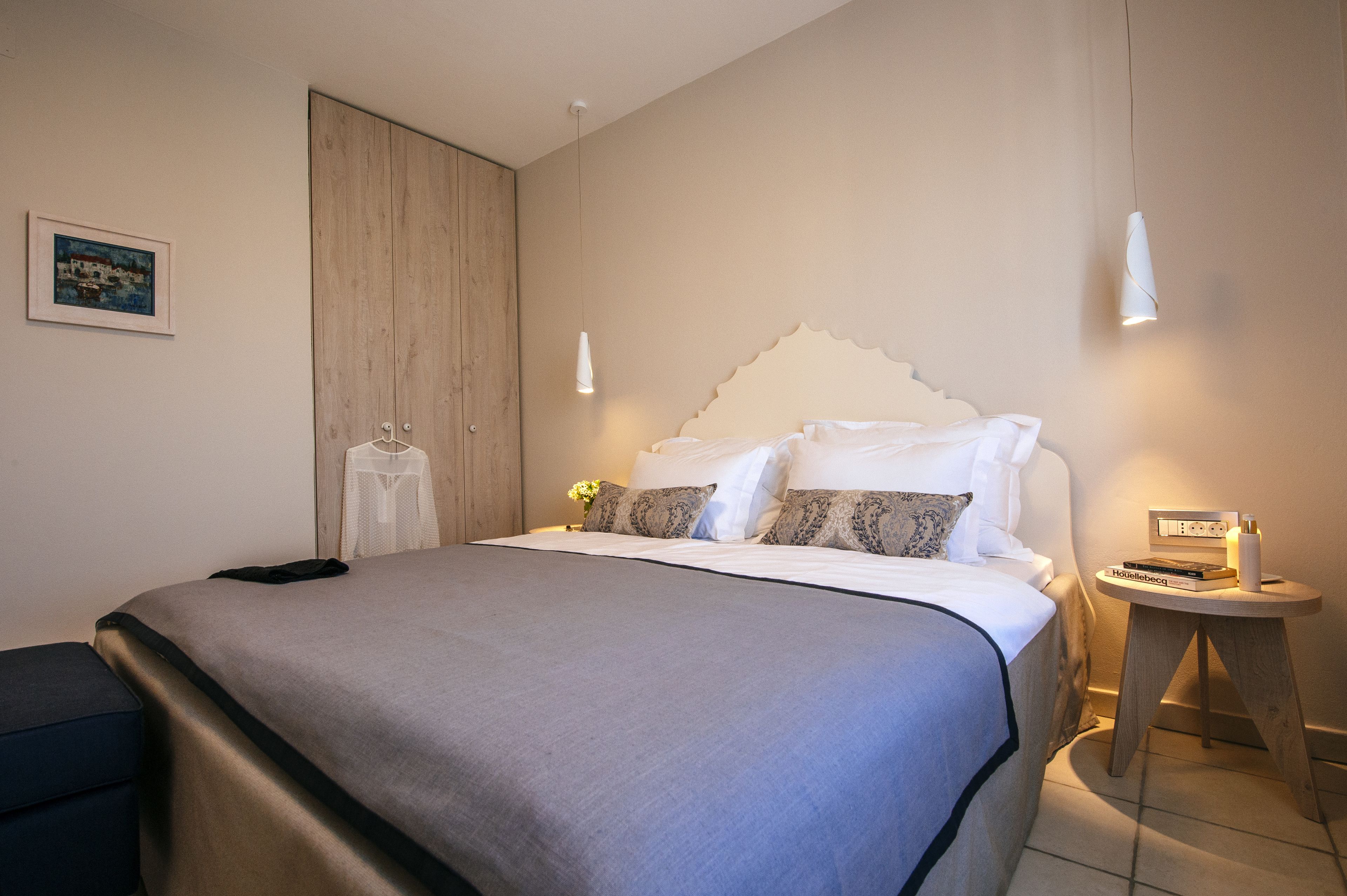 A neatly made bed with a grey bedspread, a wooden wardrobe, and a bedside table with a book and a candle.