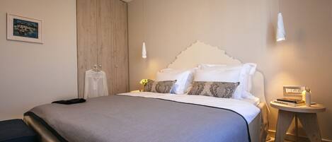 A neatly made bed with a grey bedspread, a wooden wardrobe, and a bedside table with a book and a candle.