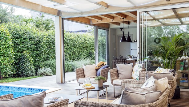 A modern outdoor living area with wicker furniture, a pool, and a glass door leading to a kitchen.