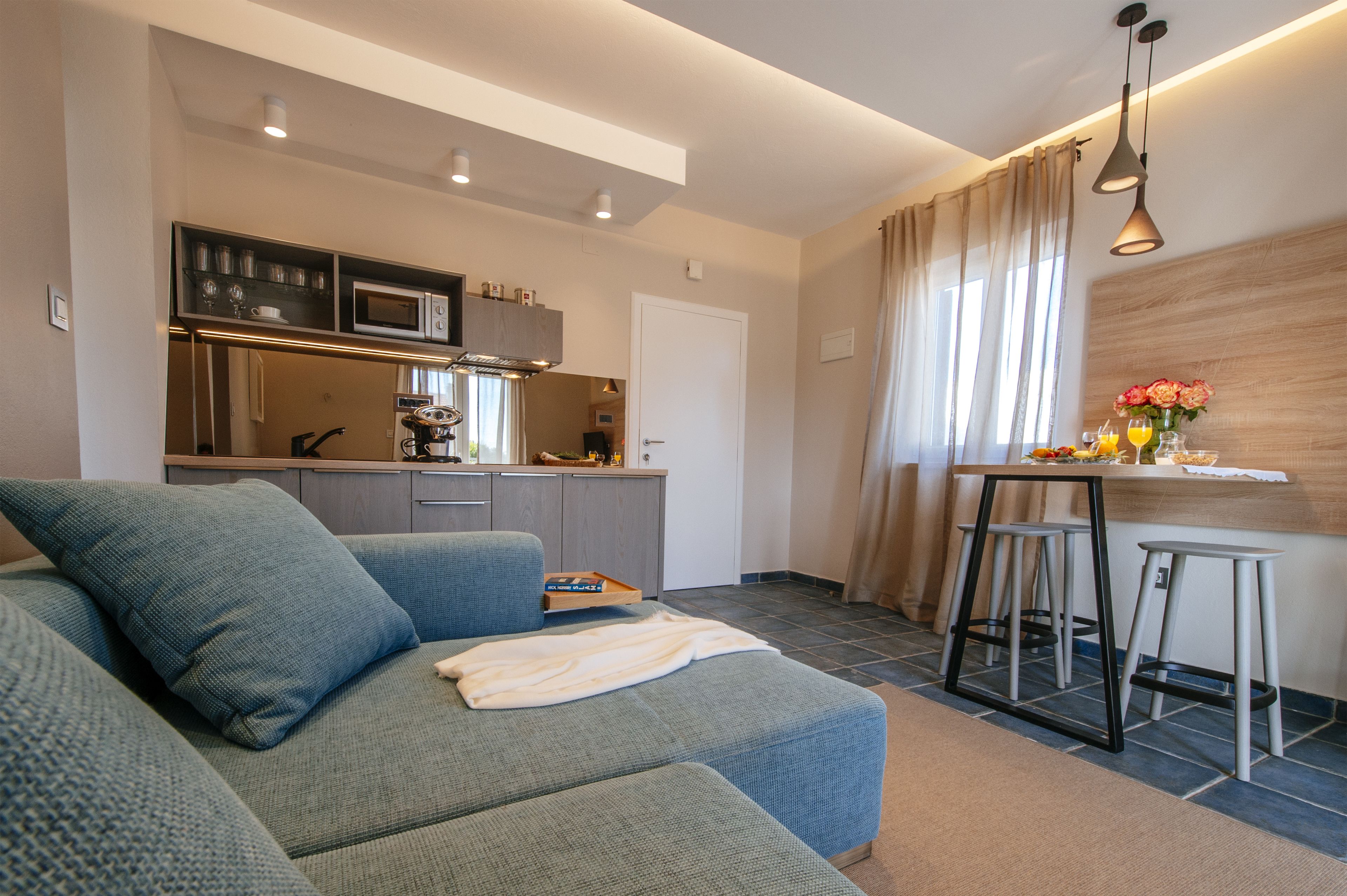 A modern living room with a sofa, a small kitchen area with a microwave and cabinets, a dining area with stools, and a window with curtains.