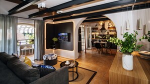 A modern living room with a sofa, a coffee table, and a TV. There is a dining area with a table and chairs. The room has wooden beams on the ceiling and a large window.