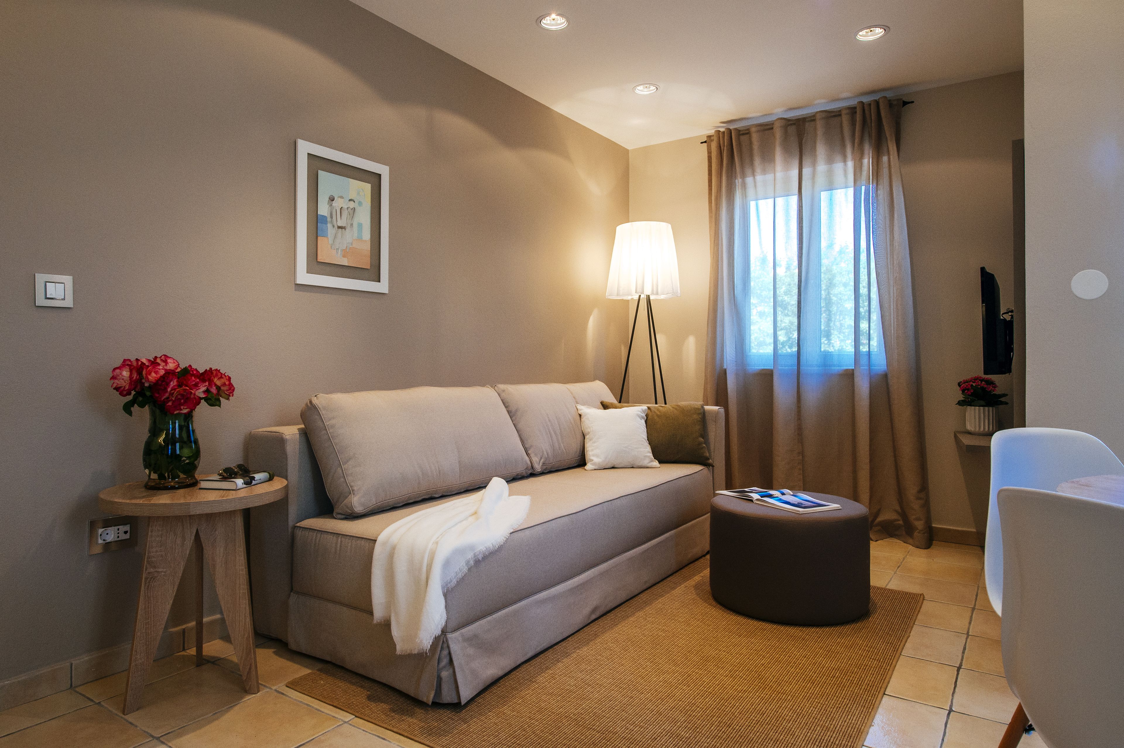Ein modernes Wohnzimmer mit einer Couch, einem Beistelltisch mit Blumenvase, einer Lampe und Blick ins Freie durch ein Fenster mit Vorhängen.