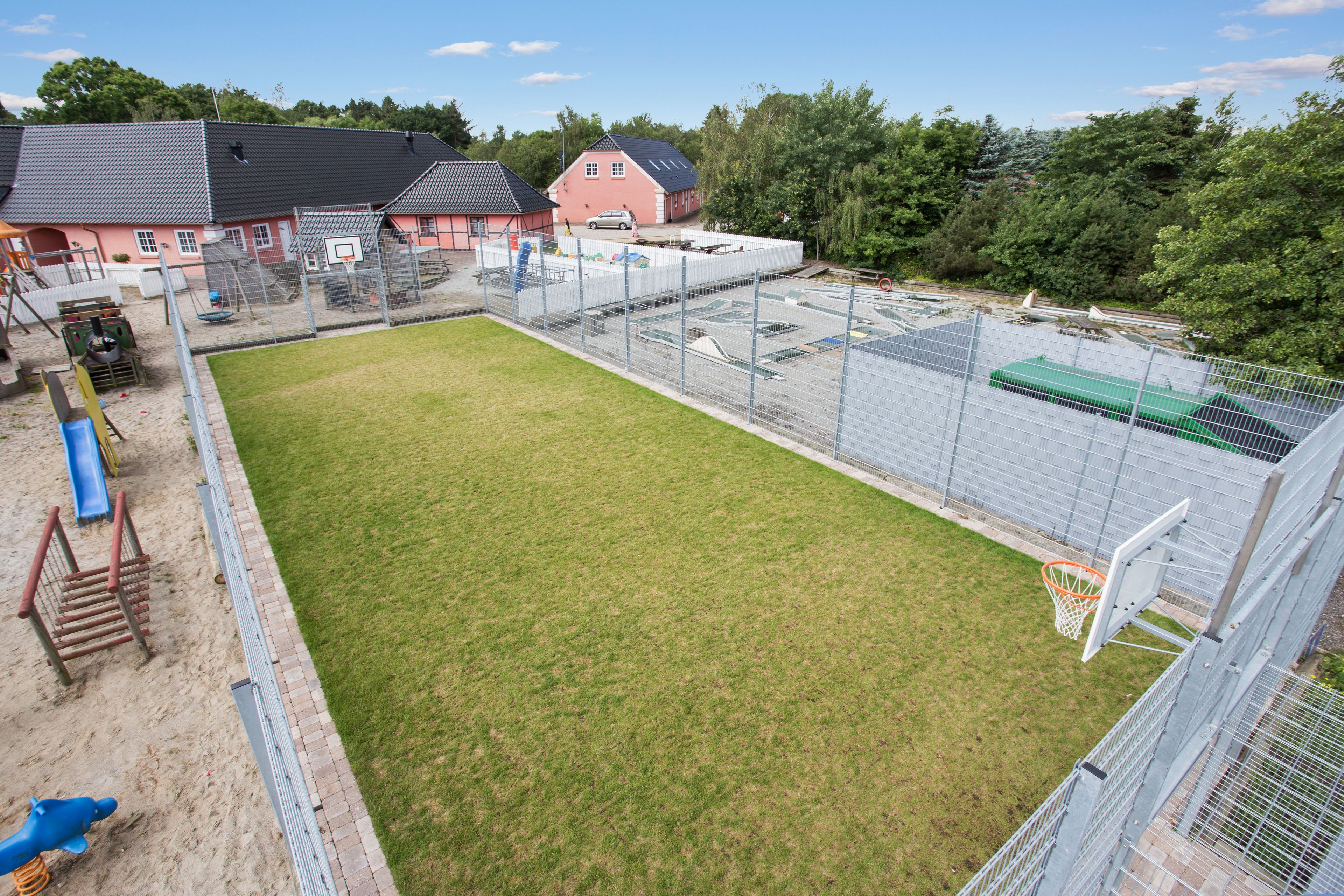 Een omheind sportterrein met een basketbalring, een speeltuin met glijbanen, en een gebouw met een rood dak.