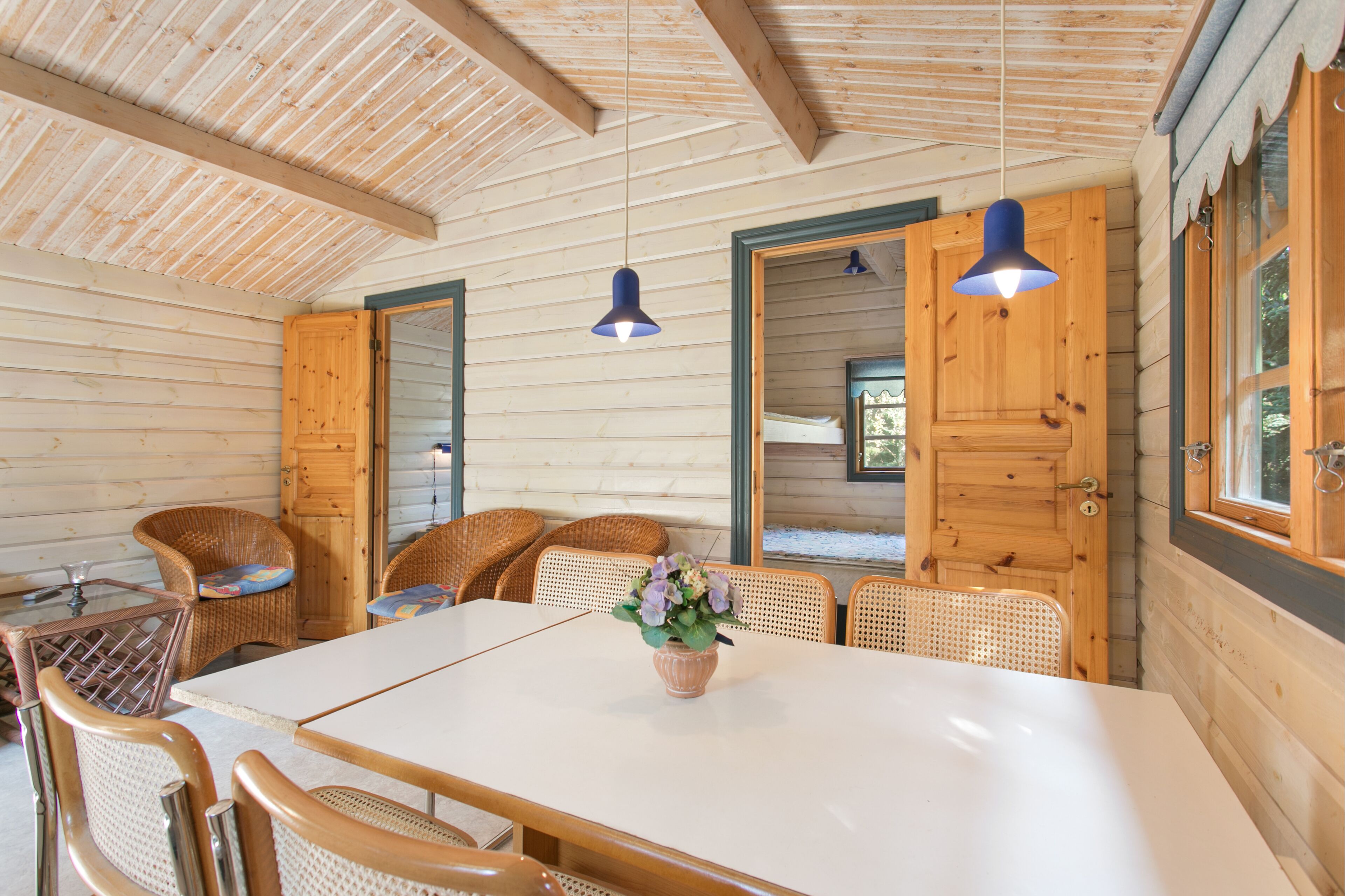 Interior de una cabaña de madera con una mesa de comedor, sillas de mimbre y un florero con flores.