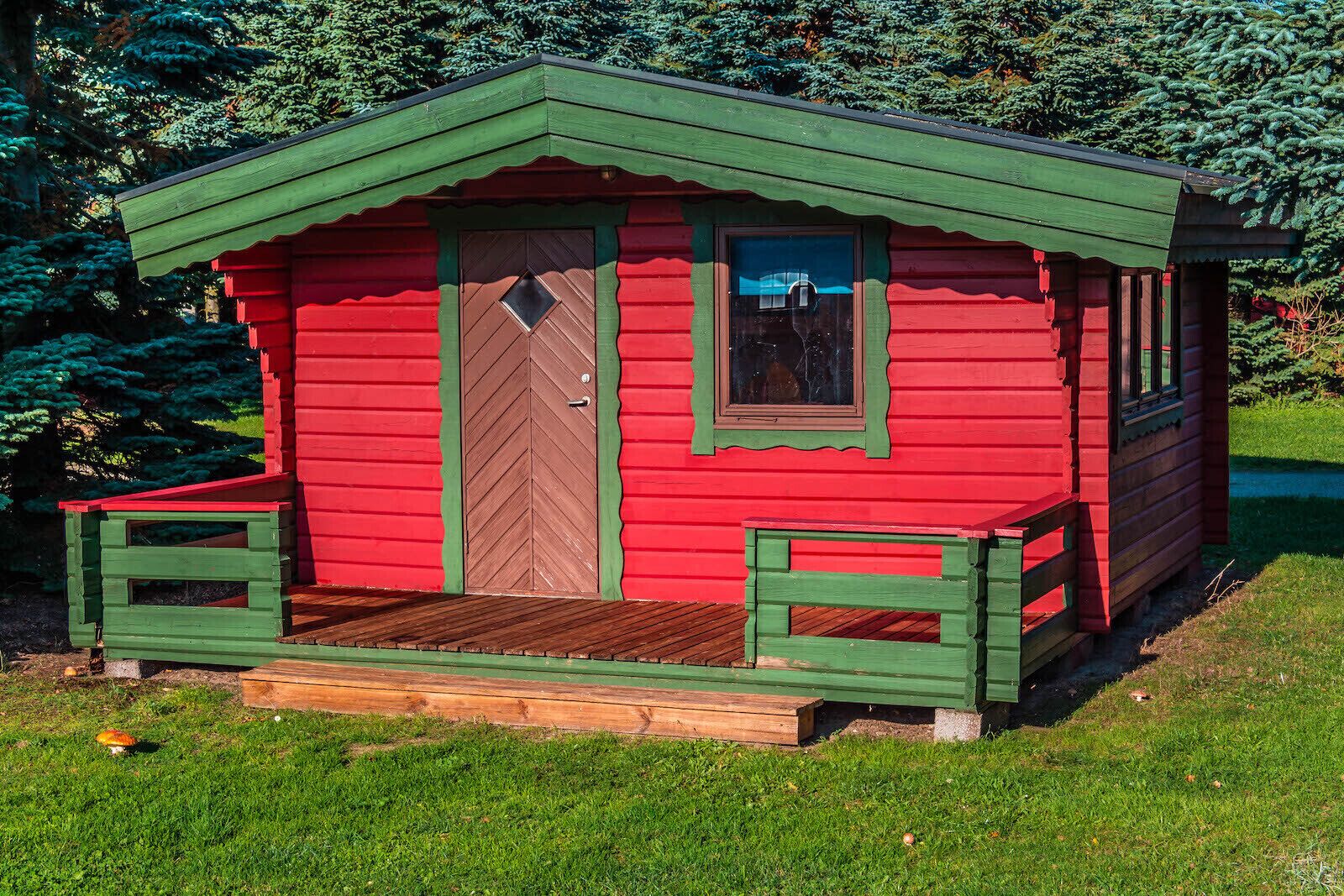 A small, red wooden cabin with a green roof, a wooden door, and a window.