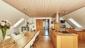 A modern kitchen with wooden cabinetry and a central island, a dining area with a long table and chairs, and a view of the adjacent living space through an open door. - Kongernes feriepark Camping (Graested)
