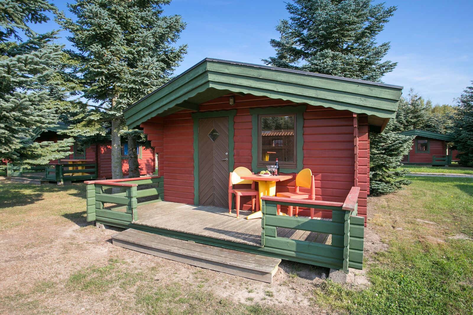 Une petite cabane à un seul étage, dotée d’un porche vert et d’une table avec des chaises.
