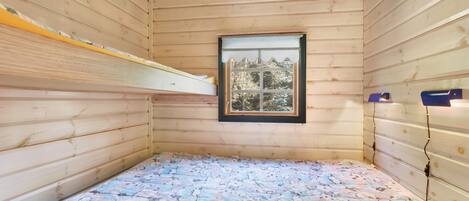 A small wooden cabin room with a bunk bed, a window with a view of trees, and a bed with a patterned blanket.