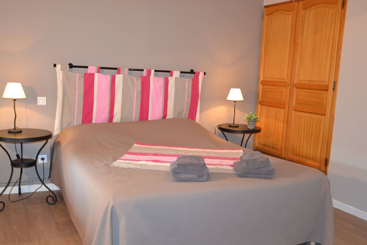 A neatly made bed with a striped bedspread, two bedside tables with lamps, a wooden wardrobe, and a potted plant on one of the tables.