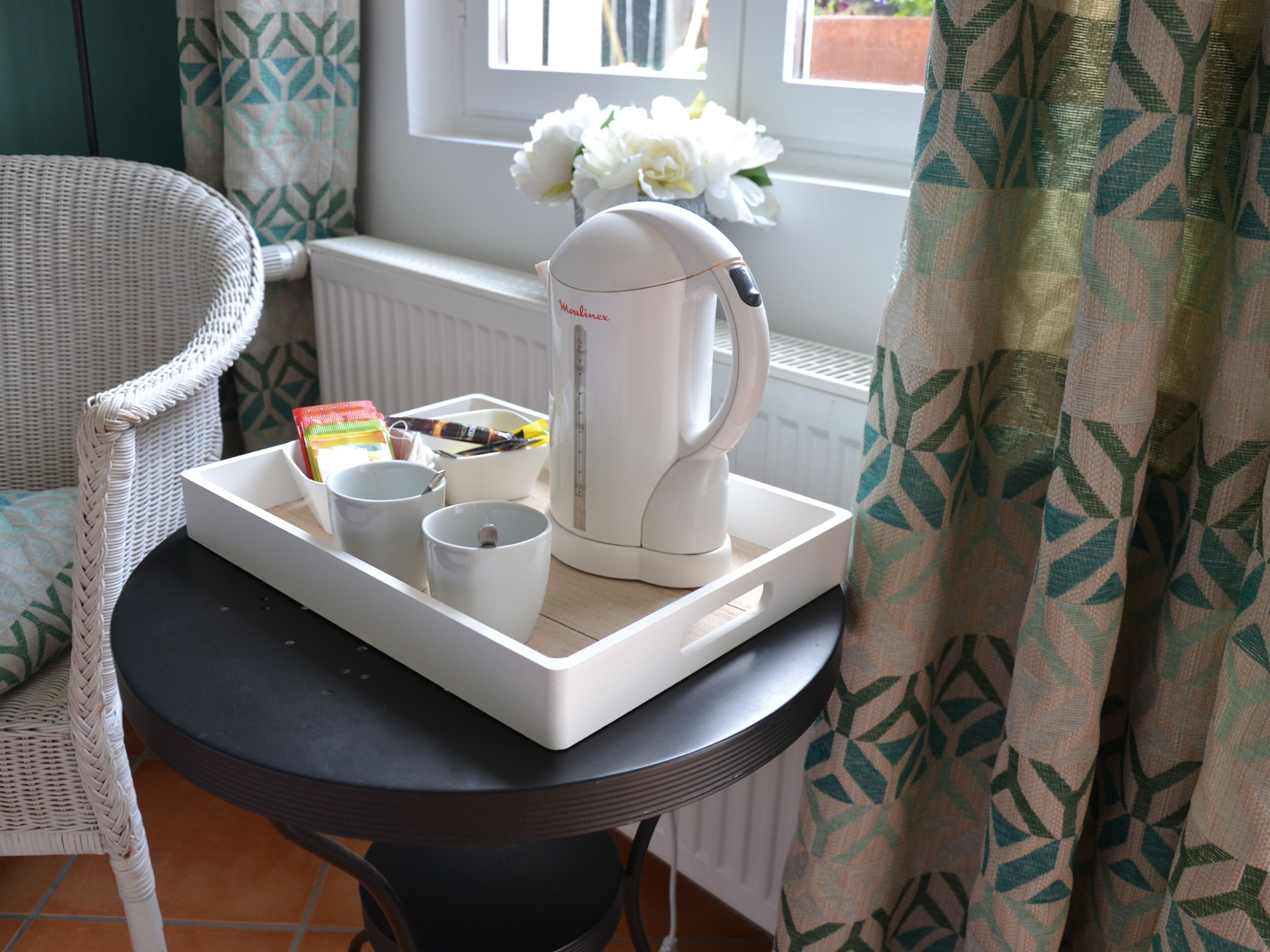 A tray with a coffee maker, cups, and a box of tea bags on a table near a window with curtains.
