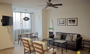 A living room with a sofa, two armchairs, a coffee table, a TV mounted on the wall, and a ceiling fan. There are two framed pictures on the wall and a surfboard leaning against the wall. - "Tortuga",  one bedroom apartment. You can walk to Beach and Restaurants (Punta de Mita)