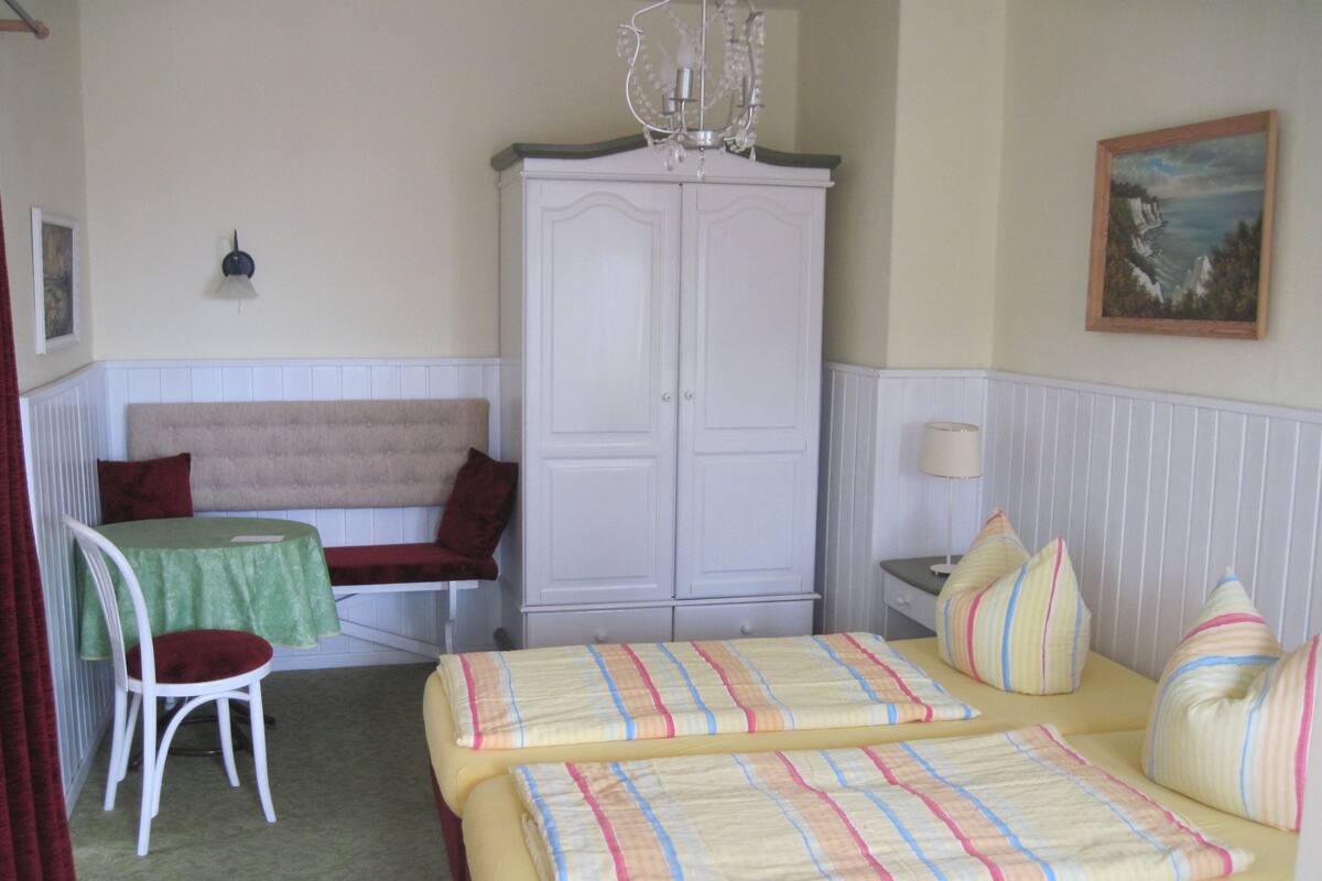 Een kamer met twee bedden, een stoel, een tafel, een kledingkast en een schilderij aan de muur.