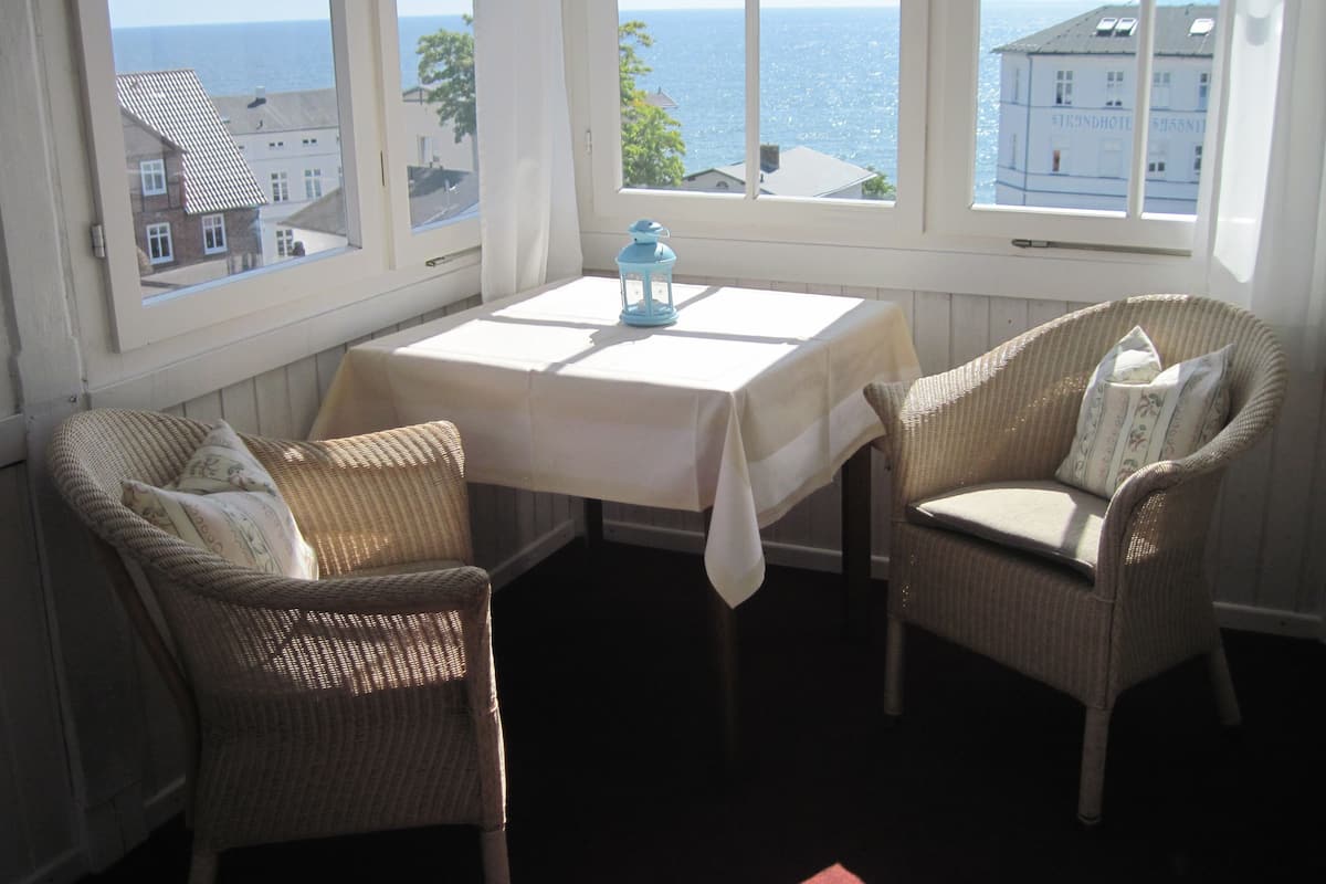 Een hoekzaal met twee rieten stoelen, een tafel met een wit tafelkleed, en uitzicht op zee door grote ramen.