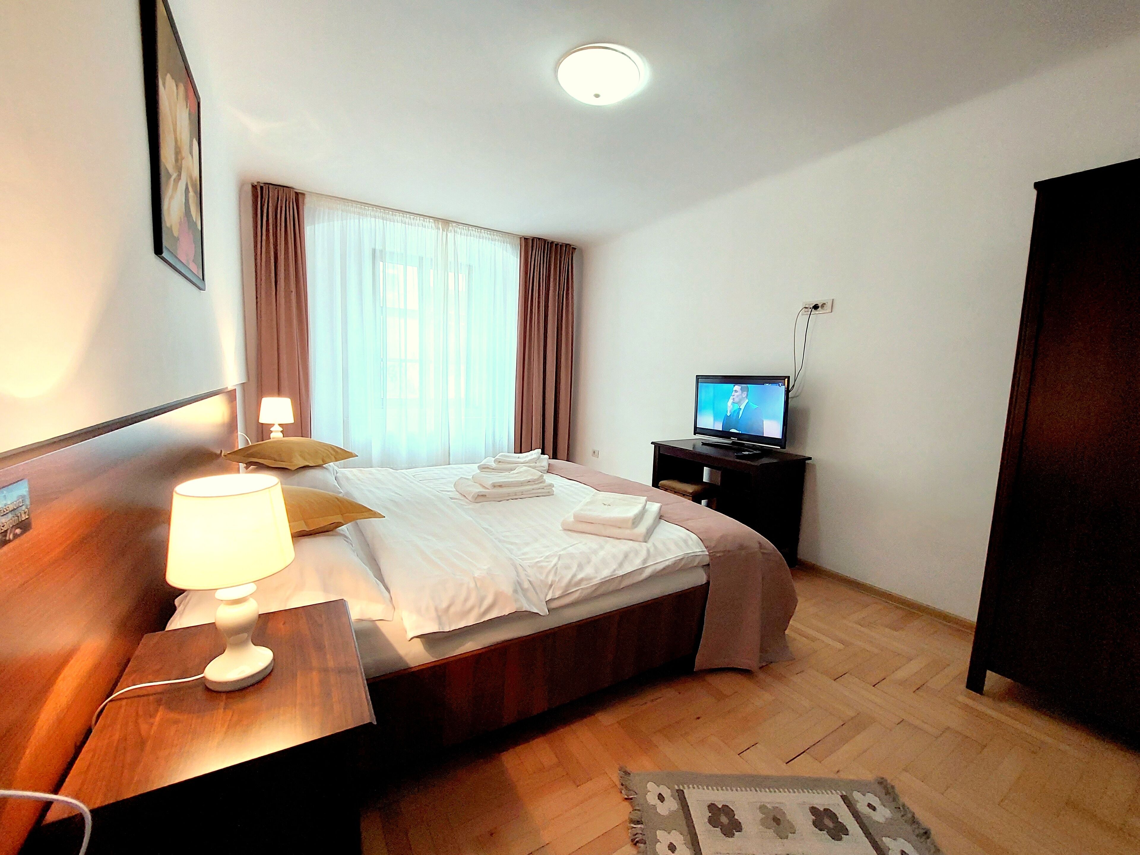 Una habitación de hotel con cama, mesita de noche con lámpara, televisión y suelo de madera.