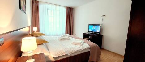 Una habitación de hotel con cama, mesita de noche con lámpara, televisión y suelo de madera.