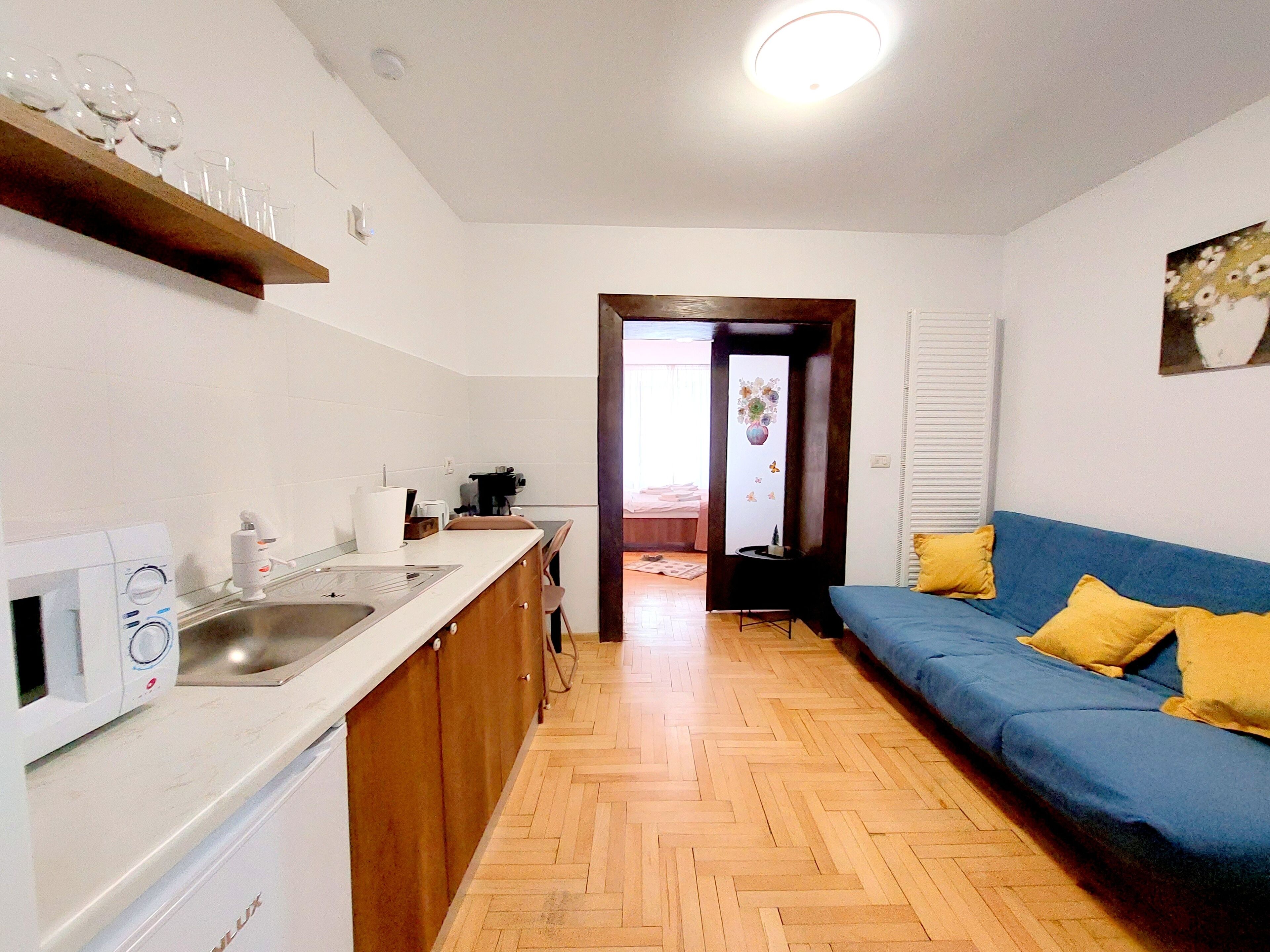 Una cucina compatta con forno a microonde, lavandino e mobili in legno. Nell'area living, un divano blu con cuscini gialli e un quadro appeso al muro.