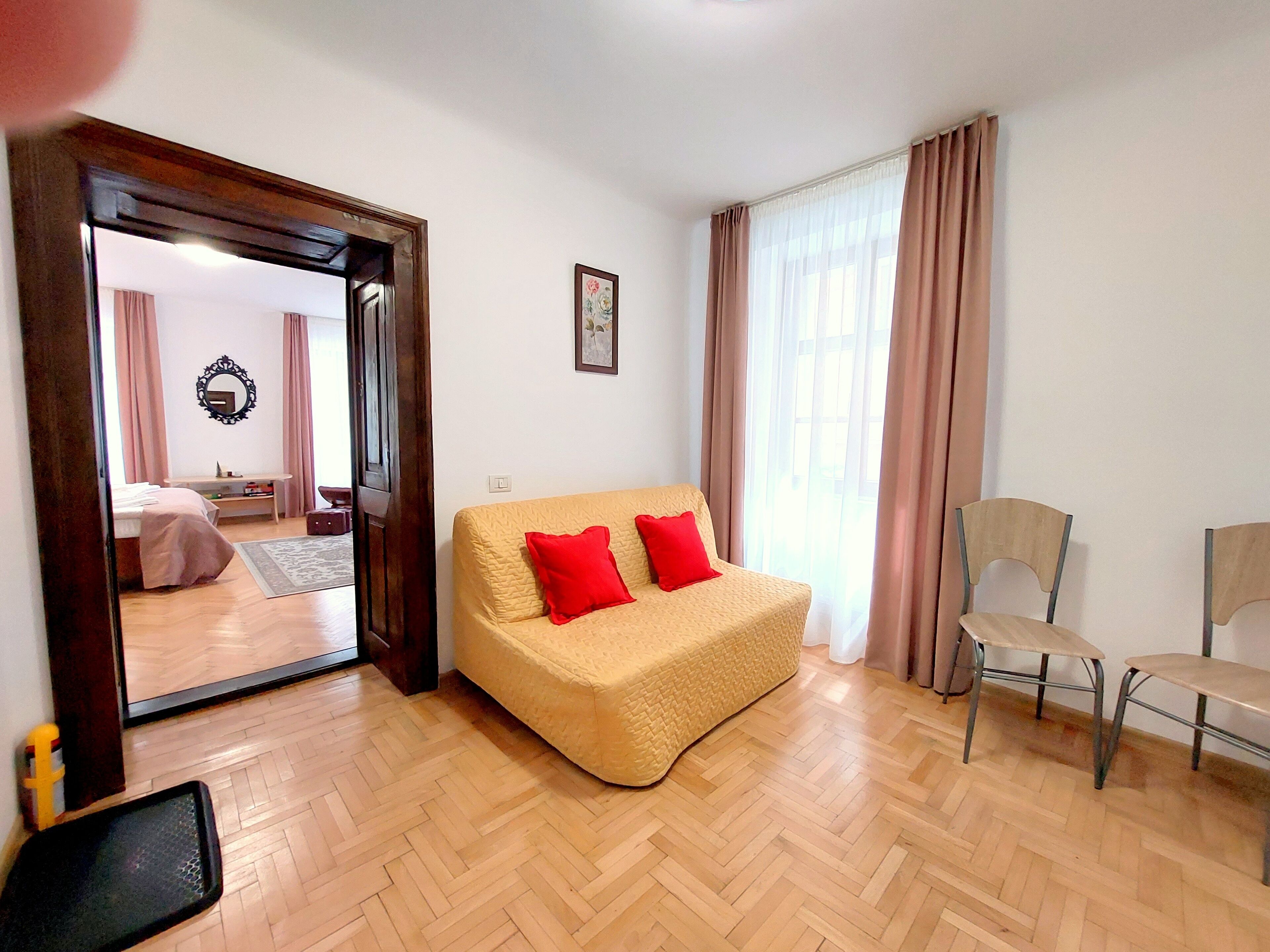 A room with a beige sofa, two red cushions, a wooden floor, a chair, and a mirror on the wall.