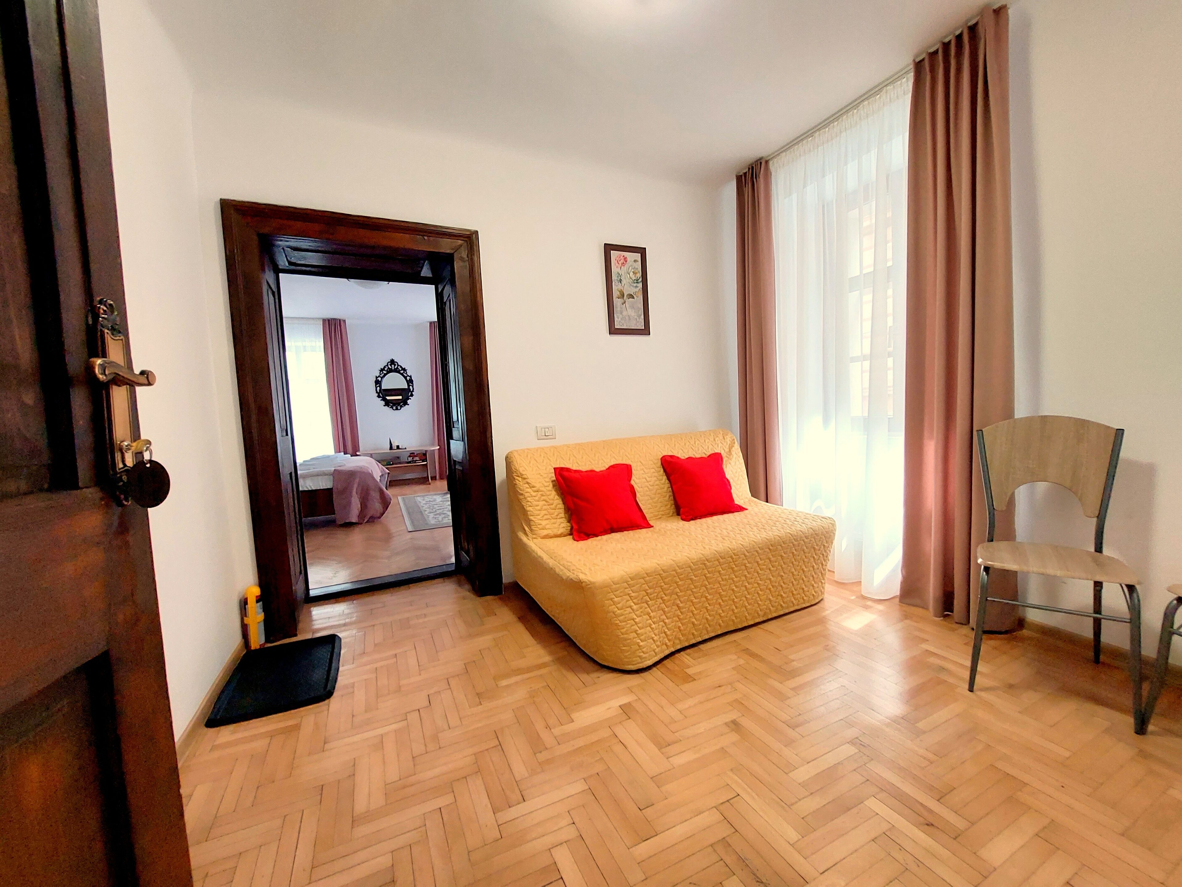 A room with a wooden herringbone floor, a beige sofa with red cushions, a wooden chair, a door with a keyhole, and a mirror on the wall.