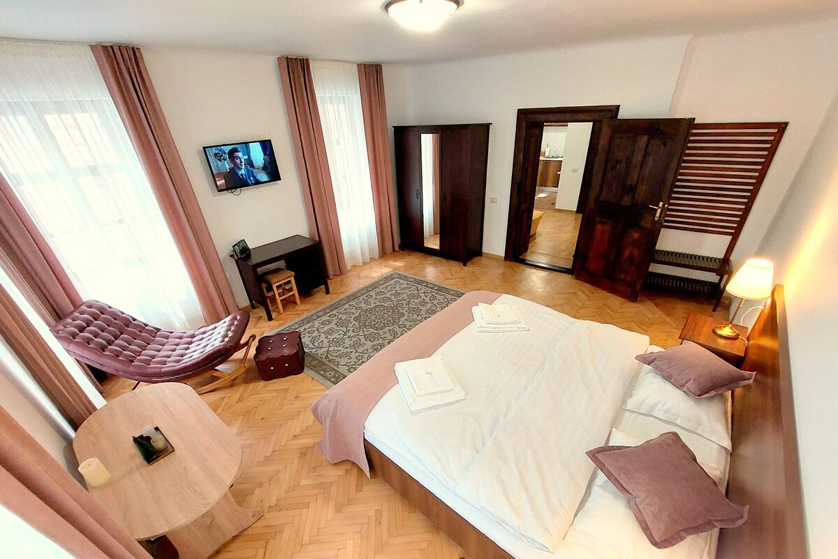 A spacious hotel room with a large bed, a sitting area with a chair, a television, and a wooden floor.