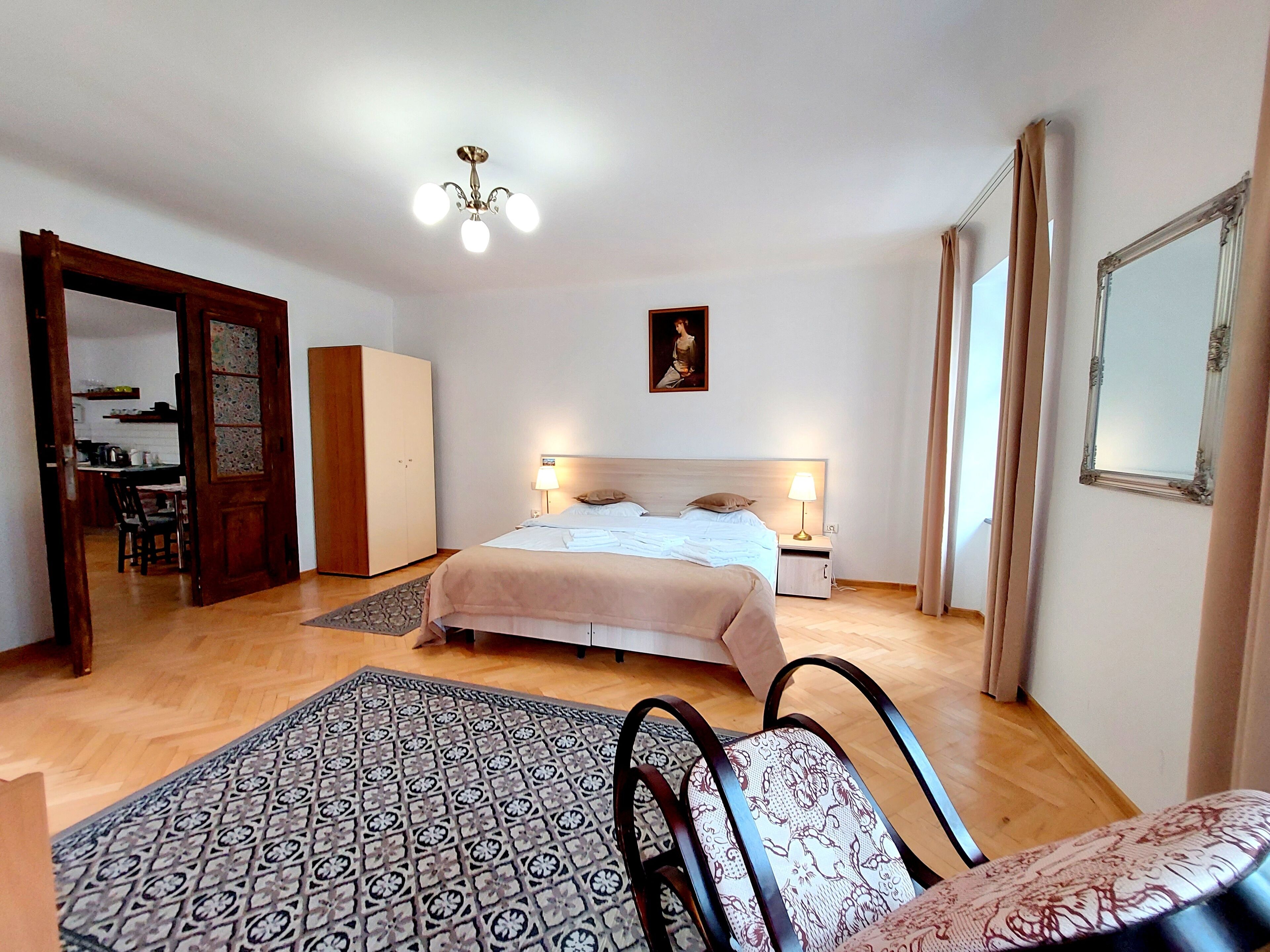 A bedroom with a bed, bedside tables, a chandelier, a wardrobe, a mirror, and a patterned rug.