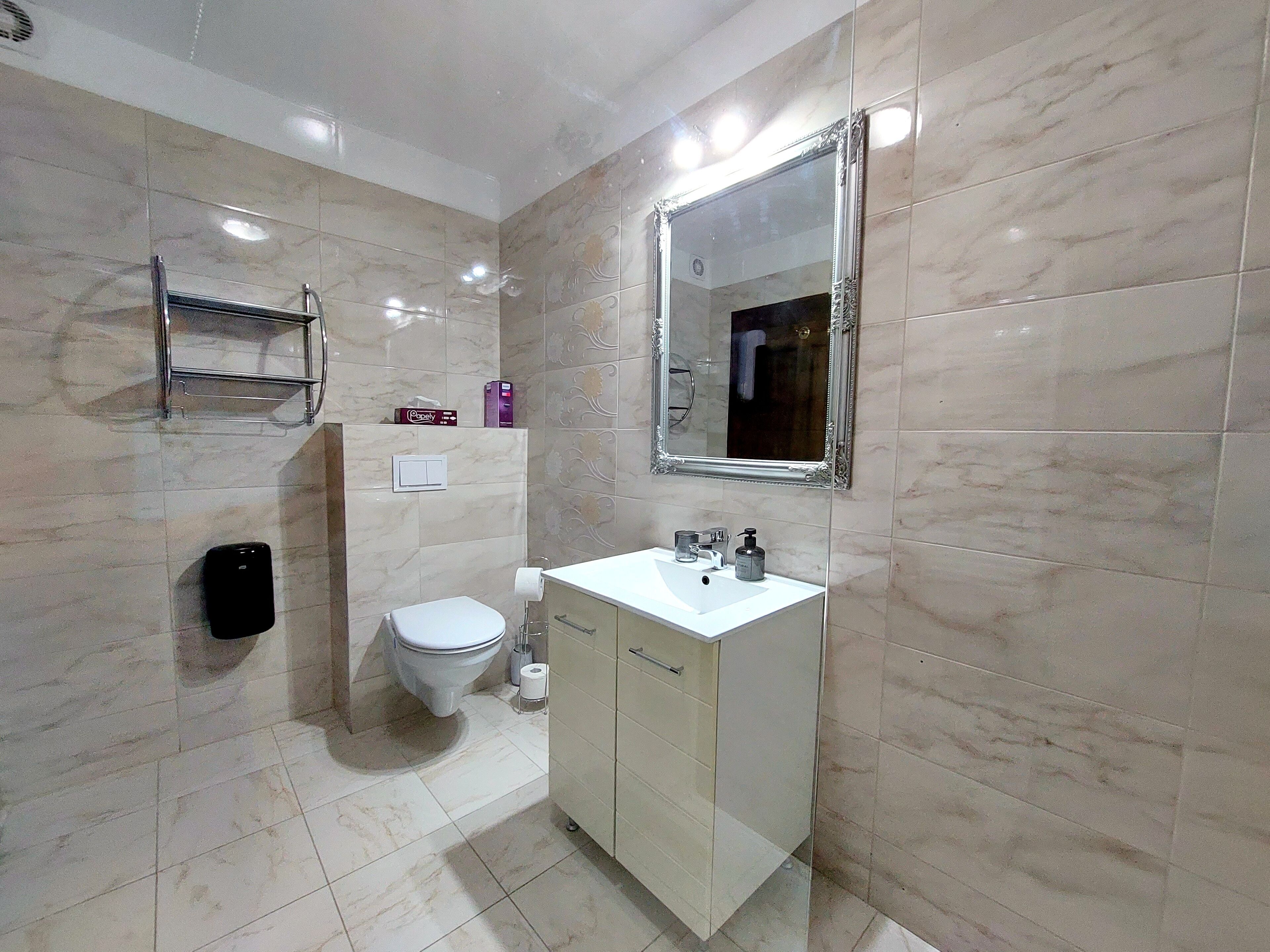 A bathroom with a toilet, sink, and mirror. There is a shelf above the toilet and a hand dryer on the wall.