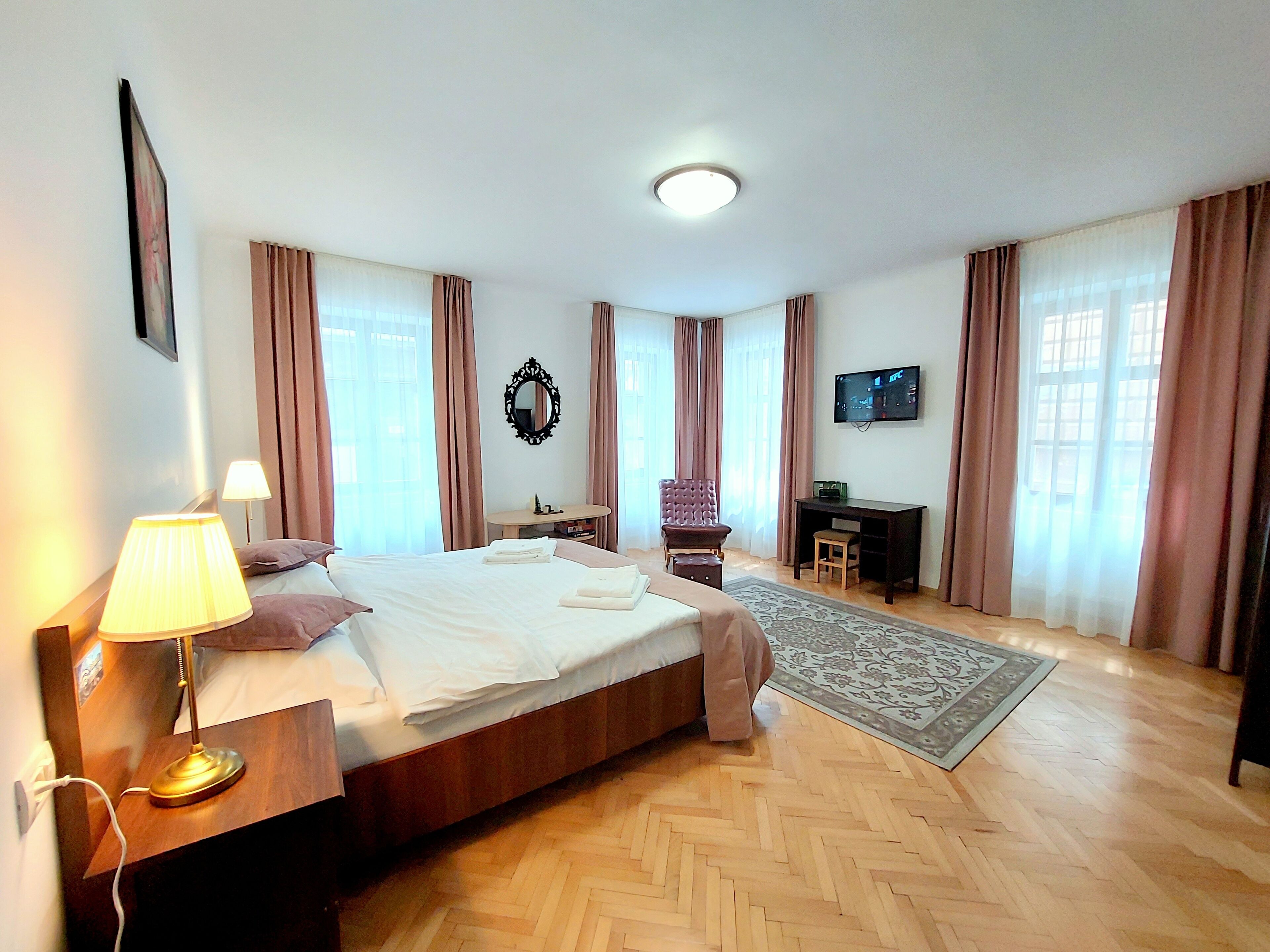 A hotel room with a bed, bedside lamp, desk, chair, TV, and a large window with curtains.