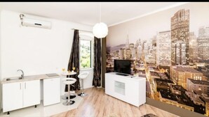 Quarto moderno com mural da paisagem urbana, um suporte branco para TV, uma pequena cozinha e uma janela com cortinas.