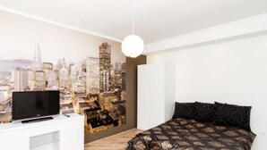 Quarto com um mural da paisagem urbana, uma cama com roupa de cama estampada, uma TV em uma cômoda branca e um lustre.