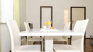 Uma área de refeições com uma mesa e cadeiras brancas, uma mesa de café da manhã com croissants e suco de laranja, e duas portas ao fundo.