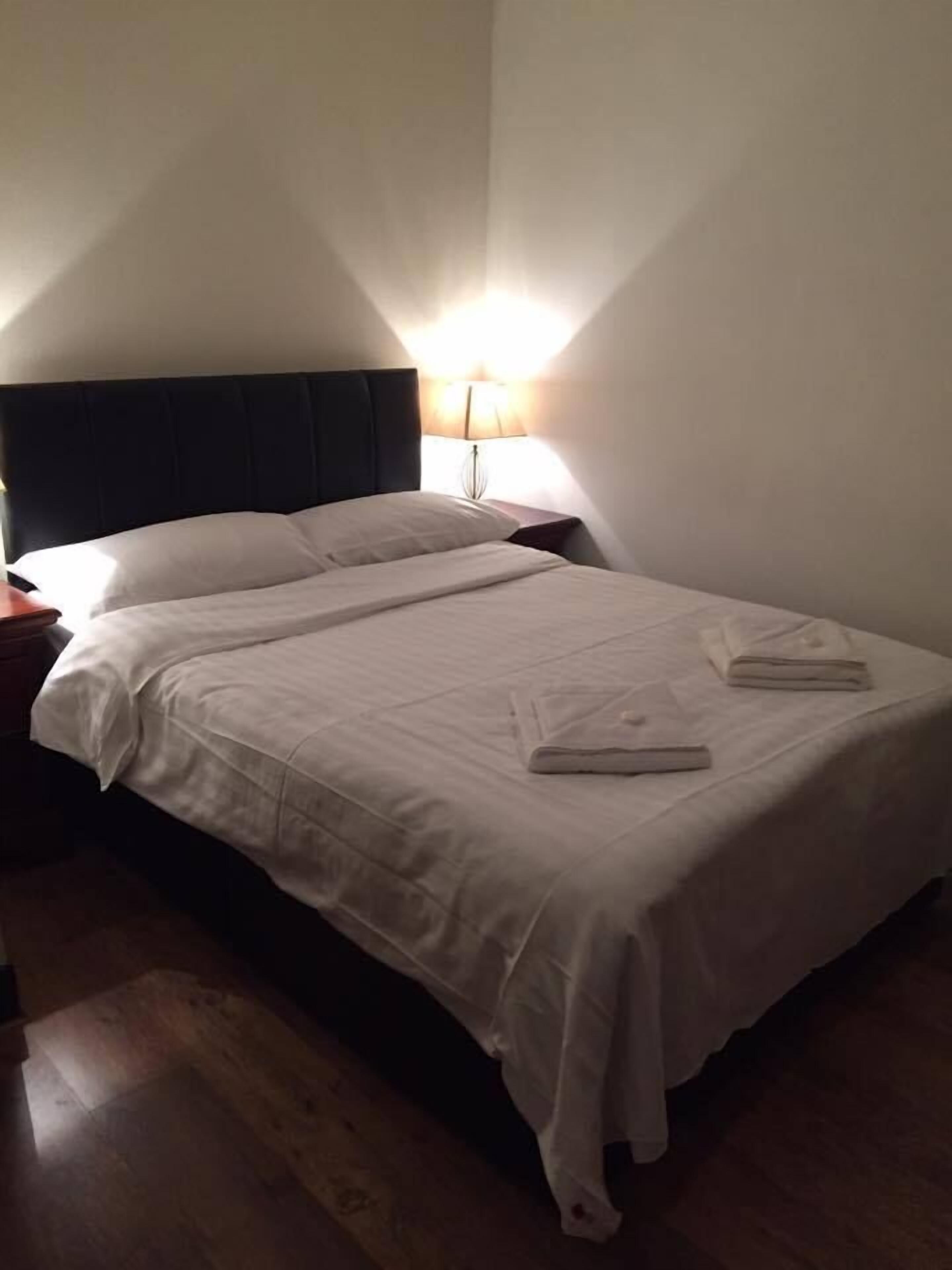 Una cama bien hecha con sábanas blancas, cabecera oscura y lámpara de noche.