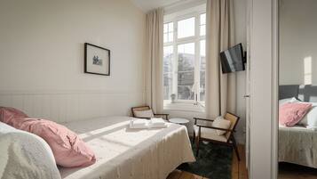 Une chambre à coucher avec un lit, une chaise, une petite table, un téléviseur et une fenêtre avec des rideaux.