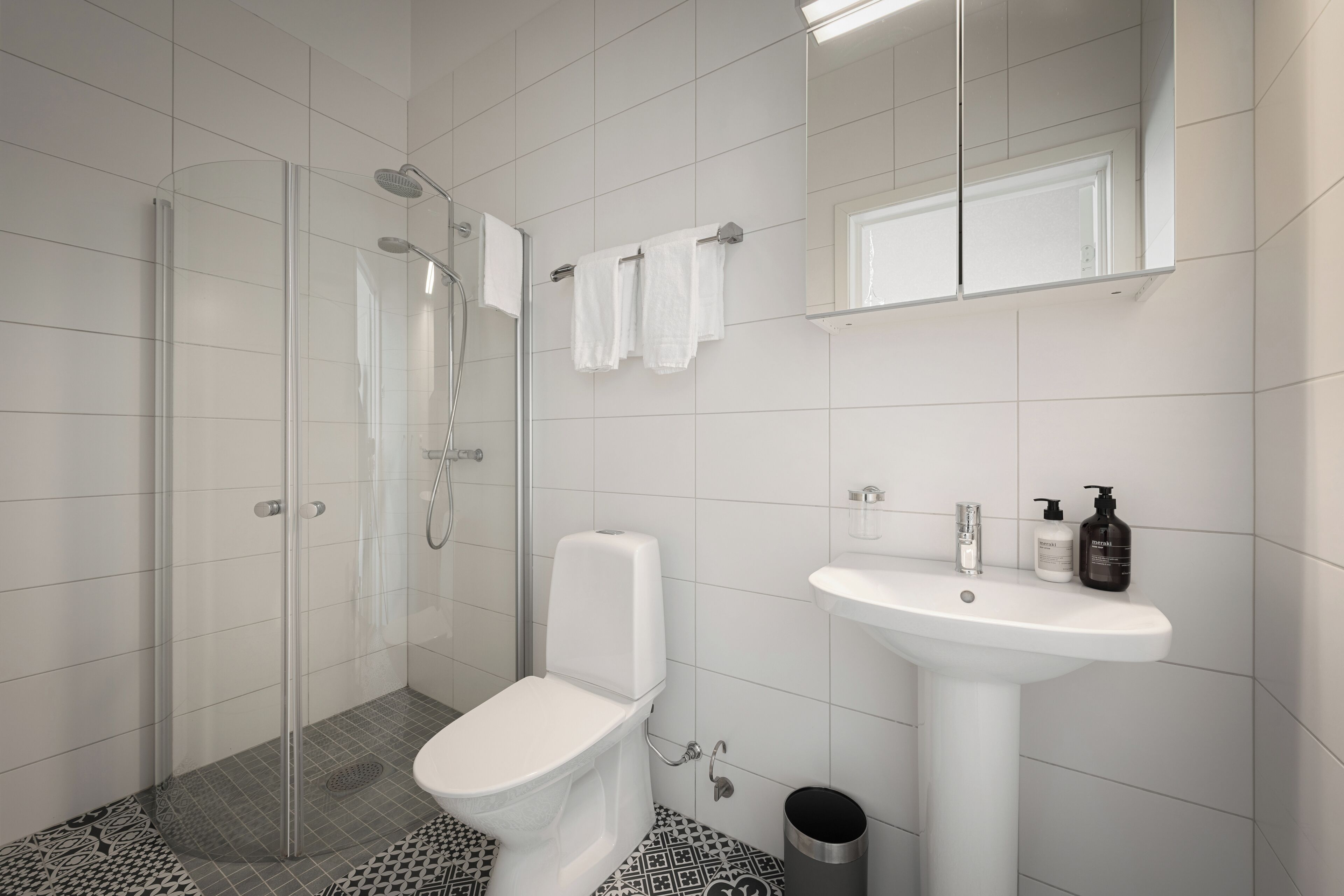 Un baño moderno con ducha de vidrio, inodoro blanco, lavamanos con artículos de aseo y espejo sobre el lavamanos.