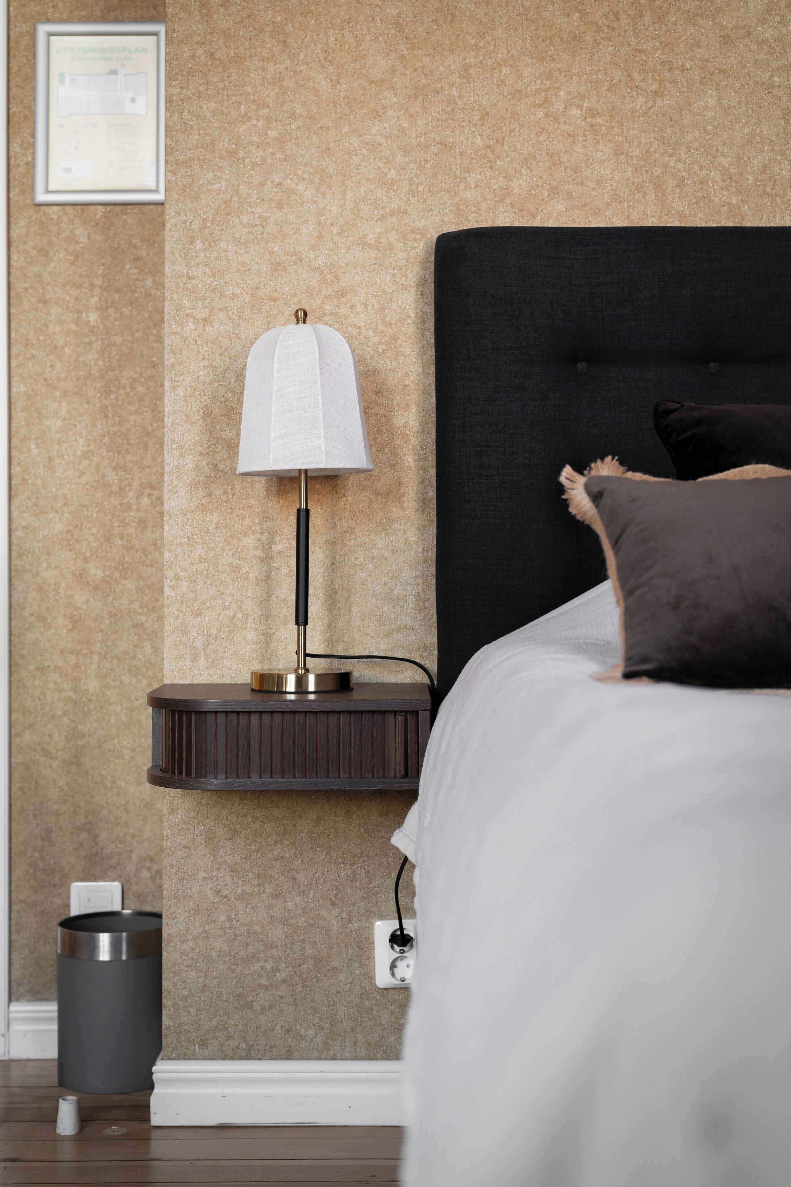 A neatly made bed with a black headboard, a bedside lamp, a trash can, and a power outlet on the wall.