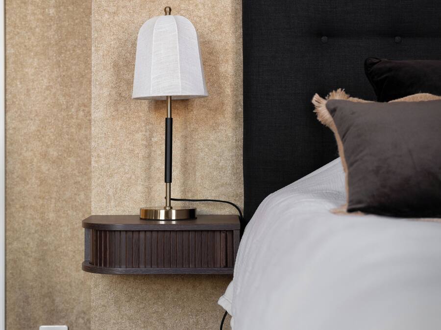 A neatly made bed with a black headboard, a bedside lamp, a trash can, and a power outlet on the wall.