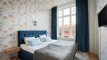 A bedroom with a blue headboard, a bed with grey bedding, a wall-mounted TV, and a window with blue curtains.