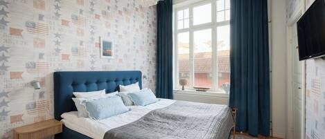 A bedroom with a blue headboard, a bed with grey bedding, a wall-mounted TV, and a window with blue curtains.