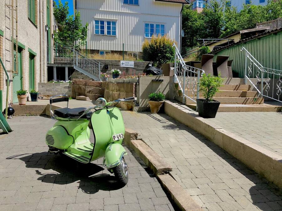 Una scooter verde antigua estacionada en una calle empedrada con un muro de piedra y escalones que conducen a un edificio.